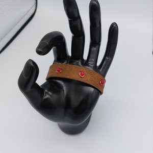 May include: A brown leather bracelet with three pink gemstones. The bracelet is on a black hand model.