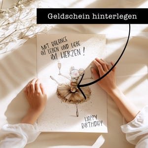 May include: A white card with a hand-drawn illustration of a mouse wearing a tutu and holding a glass of champagne. The text "Mit Balance im Leben und Liebe im Herzen!" is written in black at the top of the card. The text "Happy Birthday" is written in black at the bottom of the card. The text "Geldschein hinterlegen" is written in black on a black rectangle at the top of the image.