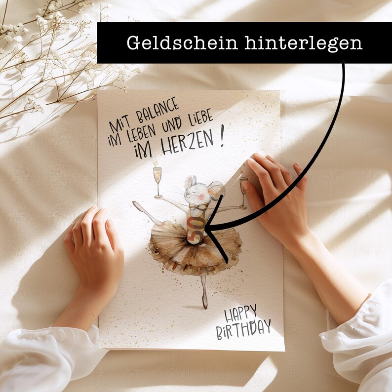 May include: A white card with a hand-drawn illustration of a mouse wearing a tutu and holding a glass of champagne. The text "Mit Balance im Leben und Liebe im Herzen!" is written in black at the top of the card. The text "Happy Birthday" is written in black at the bottom of the card. The text "Geldschein hinterlegen" is written in black on a black rectangle at the top of the image.