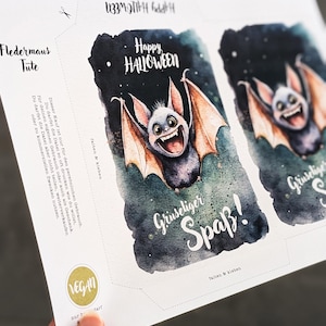 May include: Printable Halloween party favor bags featuring a cute cartoon bat with the text "Happy Halloween" and "Gruseliger Spa&szlig;!" (German for "Spooky Fun!"). The bags are designed to be folded and glued together.