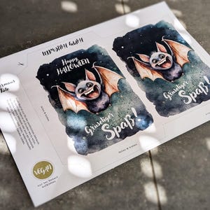 May include: Two printable Halloween treat bags featuring a cute cartoon bat with a toothy grin. The bags are decorated with a starry night sky background and the text "Happy Halloween" and "Gruseliger Spa&szlig;!" which translates to "Spooky Fun!" in English.