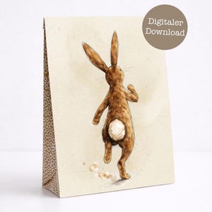 Puede incluir: Una bolsa de regalo de papel marrón con una ilustración de acuarela de un conejo corriendo. El conejo es marrón con una cola blanca. La bolsa tiene un diseño estampado en el lateral y el texto "Digitaler Download" en un círculo marrón.