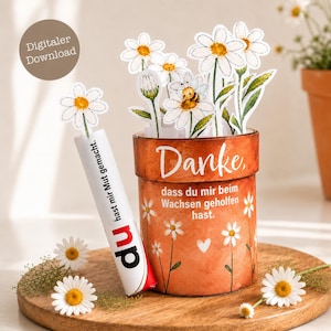 Könnte beinhalten: Ein zylindrischer orangefarbener Behälter mit weißen Gänseblümchen-Illustrationen und dem Text "Danke, dass du mir beim Wachsen geholfen hast." Daneben steht eine weiße Röhre mit dem Text "du hast mir Mut gemacht" und einem Gänseblümchen.