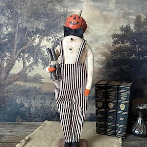 May include: A fabric doll featuring a carved orange pumpkin head with a black jack-o'-lantern face, wearing a black bow tie, cream-colored shirt with two buttons, and black and white striped overalls. The doll holds a bundle of small wooden sticks.