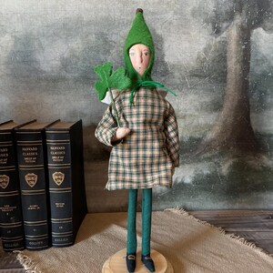 May include: A handcrafted St. Patrick's Day doll. The doll wears a green pointed hat, a plaid dress, and holds a green felt shamrock. It stands on a wooden base with green legs and black shoes. The background includes vintage books.