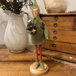 May include: A handmade folk art doll with a green tunic and hat, holding a brown mushroom with white spots. The doll has orange legs and green boots, standing on a wooden base. The background includes a white pitcher and a wooden cabinet.