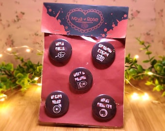 Puede incluir: Cinco pines de botón negros y blancos con texto rojo. Los pines están empaquetados en una bolsa de plástico transparente con una tarjeta de encabezado roja y negra que dice "Nova v Rose". Los pines presentan el siguiente texto: "Wild Child", "Shadow Queen", "Ugly & Proud", "Bicorn Nerd" y "Meat Crafter".
