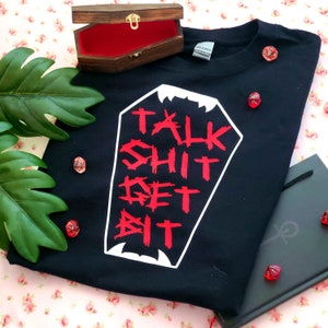 May include: Black t-shirt with a white coffin graphic and the text "Talk Shit Get Bit" in red.