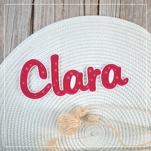 Könnte beinhalten: Ein personalisierter Namensaufnäher mit dem Namen Clara in einer kräftigen, dunkelrosa Schriftart mit hellrosa Punkten. Die Buchstaben sind mit weißem Garn umrandet und haben eine erhabene, gestickte Textur für ein klares Design.