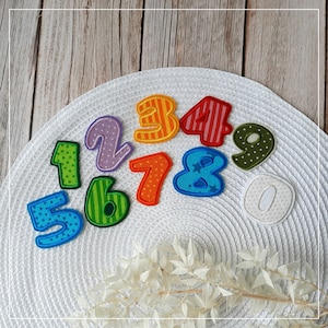 Könnte beinhalten: Buntfarbene Filzpatches in Form von Zahlen von 1 bis 9 und 0. Die Patches sind in einem Kreis auf weißem Hintergrund angeordnet.