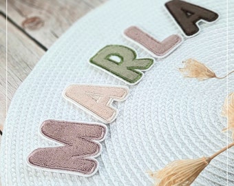 May include: The name MARLA spelled out in individual fabric letters with a textured surface. The letters are colored in shades of dusty rose, light beige, sage green, and dark brown, each outlined in white, arranged on a white woven circular mat.