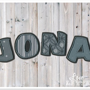 Könnte beinhalten: Der Name JONA, buchstabiert in vier einzelnen Stoffbuchstaben. Jeder Buchstabe hat einen dunkelgrauen Rand und ein einzigartiges Muster: das J hat kleine Sterne, das O vertikale Streifen, das N ein florales geometrisches Muster und das A ist grau.
