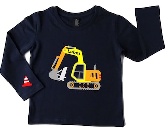 May include: A navy blue long-sleeve shirt featuring a yellow and orange excavator graphic with the name Lukas printed on the arm of the machine. A small red and white traffic cone graphic is positioned on the left sleeve of the shirt.