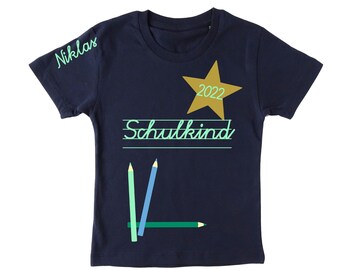May include: A navy blue t-shirt with the text "Niklas" on the left sleeve, a gold star with the text "2022" inside, and the text "Schulkind" in green cursive writing. Three colored pencils are shown below the text.