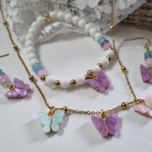 Könnte beinhalten: Ein Schmuckset bestehend aus einer goldfarbenen Kettenhalskette mit drei Schmetterlingsanhängern, einem Stretch-Armband mit weißen Perlen und einem Schmetterlingsanhänger sowie zwei passenden Ohrhängern mit bunten Perlen.