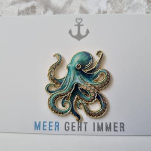 Könnte beinhalten: Eine goldfarbene Oktopus-Anstecknadel mit blau-türkisen Emaille-Details, präsentiert auf einer weißen Karte mit einem grauen Anker-Symbol oben und dem in Blau gedruckten Text MEER GEHT IMMER am unteren Rand der Karte.