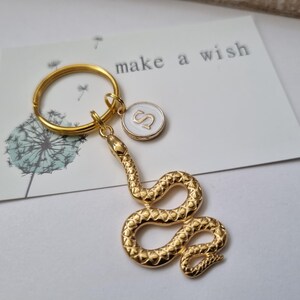 Könnte beinhalten: Ein goldfarbener Schlangen-Schlüsselanhänger mit einem runden Anhänger mit dem Buchstaben "S" auf weißem Hintergrund. Der Schlüsselanhänger ist an einer "make a wish"-Karte mit einer Pusteblumen-Illustration befestigt.