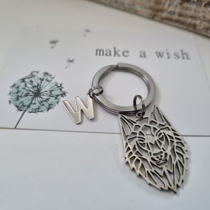Könnte beinhalten: Silberner Schlüsselanhänger mit einem Wolfskopf-Anhänger und einem Buchstaben W-Anhänger. Der Schlüsselanhänger befindet sich auf einer weißen Karte mit dem Text "make a wish".