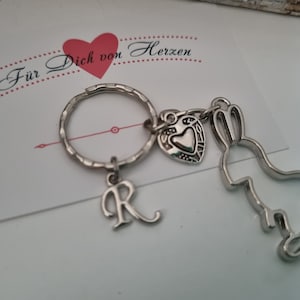 Könnte beinhalten: Silberner Schlüsselanhänger mit einem Herz-Anhänger, einem Hasen-Anhänger und einem Buchstaben R-Anhänger. Der Schlüsselanhänger ist an einem silbernen Schlüsselring befestigt. Der Text "Für Dich von Herzen" ist im Hintergrund sichtbar.
