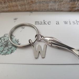 Könnte beinhalten: Ein silberfarbener Metallschlüsselanhänger mit einem runden Spaltring, einem flachen Buchstaben-Anhänger W und einem dreidimensionalen Fisch-Anhänger. Der Schlüsselanhänger liegt auf einer weißen Karte mit dem Text make a wish.