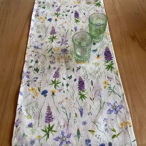 May include: A white table runner with a floral pattern featuring purple, blue, and yellow wildflowers. Two green, patterned drinking glasses sit on the runner. The runner is made of a linen-like material and is placed on a wooden table.
