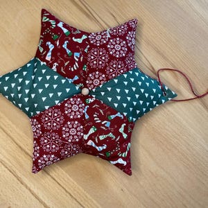 Könnte beinhalten: Ein sternförmiger Weihnachtsschmuck mit Patchwork-Design. Der Schmuck hat abwechselnde Abschnitte aus dunkelrotem Stoff mit weißen Schneeflocken- und Urlaubs-Mustern und grünem Stoff mit weißen Dreiecksmustern. In der Mitte befindet sich eine kleine Holzperle.