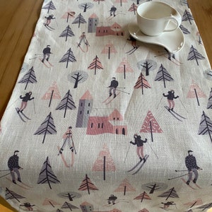 Könnte beinhalten: Cremefarbener Tischläufer mit einer Winterskiszene. Das Design zeigt Skifahrer, Bäume und Gebäude in Grau-, Rosa- und Lilatönen. Eine weiße Tasse und Untertasse liegen auf dem Läufer.