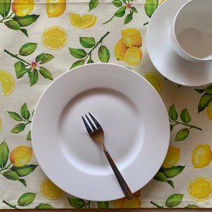 May include: A white plate, fork, teacup, and saucer set on a placemat with a lemon and leaf pattern. The placemat has a light beige background with yellow lemons and green leaves. The teacup and saucer are white, and the fork is gold.