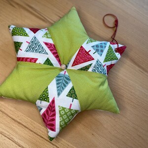 Könnte beinhalten: Ein sternförmiger Weihnachtsschmuck. Der Schmuck besteht aus grünem Stoff mit Streifenmuster und Stoff mit einem Weihnachtsbaum-Muster in Rot, Grün und Blau. In der Mitte befindet sich eine kleine Holzperle, und zum Aufhängen ist eine rote Schnur befestigt.