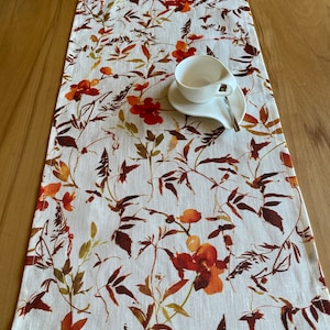 Op de afbeelding: Een witte linnen tafelloper met een bloemenpatroon in oranje, bruine en groene tinten. Een witte kop en schotel met een zilveren lepel liggen erop. De tafelloper is ongeveer 152 cm lang.