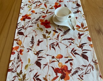 May include: A white linen table runner with a floral pattern in shades of orange, brown, and green. A white teacup and saucer with a silver spoon are placed on the runner. The runner is approximately 60 inches long.