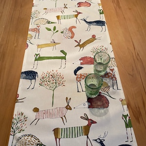Può includere: Una tovaglia color crema con una stampa di animali della foresta. Il design include volpi, cervi, scoiattoli e ricci stilizzati in vari colori. Due piccoli bicchieri verdi sono appoggiati sulla tovaglia, che misura circa 30 cm di larghezza.
