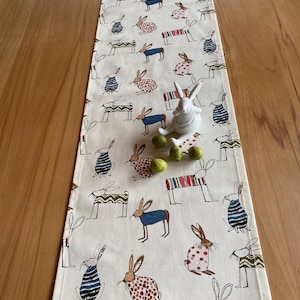 Puede incluir: Un camino de mesa de Pascua color crema con un patrón repetido de conejos ilustrados con varios atuendos. Una figura de conejo de cerámica blanca y huevos decorativos verdes están sobre el camino de mesa. El camino de mesa está sobre una mesa de madera.