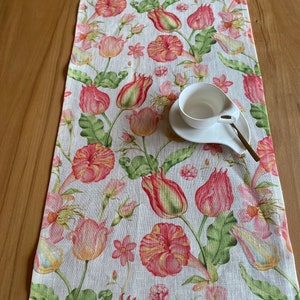 Op de afbeelding: Een bloemen tafelloper met een patroon van roze en rode tulpen en groene bladeren op een witte achtergrond. Een witte kop en schotel liggen op de loper. De loper is gemaakt van een textuurstof.