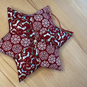 Könnte beinhalten: Ein burgunderfarbener sternförmiger Weihnachtsschmuck. Der Stoff hat ein Muster aus Schneeflocken und festlichen Socken in Weiß, Blau und Grün. Eine kleine weiße Perle und ein roter Faden sind zum Aufhängen befestigt. Auf einer Holzoberfläche.