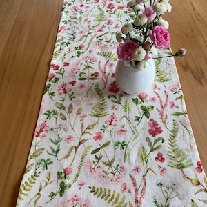 May include: A white linen table runner with a floral pattern featuring pink and red flowers and green leaves. A white vase with pink flowers sits on the runner.