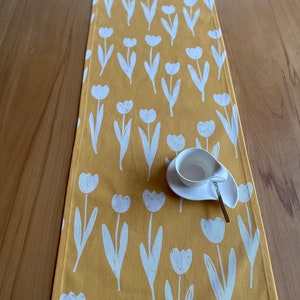 Puede incluir: Un camino de mesa amarillo con un patrón de tulipanes blancos. El camino de mesa está sobre una mesa de madera con una taza y un platillo blancos encima.