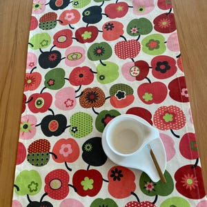 Peut inclure: Un chemin de table rectangulaire avec un motif répétitif de pommes stylisées dans des tons de rouge, rose, vert et noir sur un fond crème. Une tasse et une soucoupe en céramique blanche avec une petite cuillère dorée sont posées dessus.