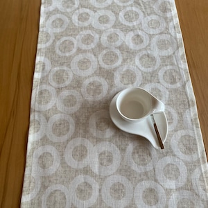 Könnte beinhalten: Beige-weiß gemusterter Tischläufer mit sich wiederholendem Kreisdesign. Eine weiße Tasse und Untertasse mit Löffelablage liegen auf dem Läufer. Der Läufer liegt auf einem Holztisch.