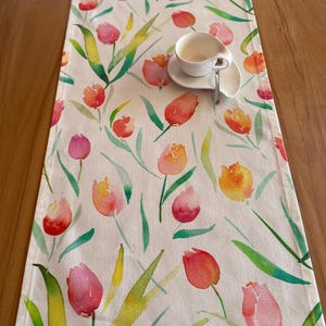 Op de afbeelding: Witte tafelloper met een aquarel tulpenontwerp in roze, oranje en geel, met groene bladeren. Een witte kop en schotel staan op de loper. Geschikt voor een eettafel.