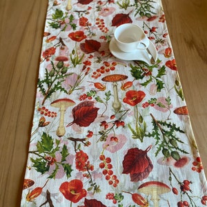 Könnte beinhalten: Ein weißer Tischläufer mit einem botanischen Druck mit roten Mohnblumen, Pilzen, Beeren und Blättern. Eine weiße Tasse und Untertasse liegen auf dem Läufer. Der Läufer besteht aus einem strukturierten Stoff.