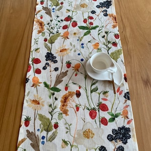May include: A white linen table runner with a floral pattern featuring red strawberries, blackberries, and yellow flowers. The runner is on a wooden table with a white cup and saucer.