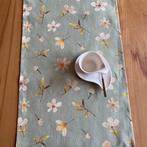 Op de afbeelding: Lichtgroene tafelloper met een bloemenpatroon van witte en gele bloemen en bruine bladeren. Een witte keramische kop en schotel liggen op de loper. De loper is gemaakt van een linnenachtig materiaal.