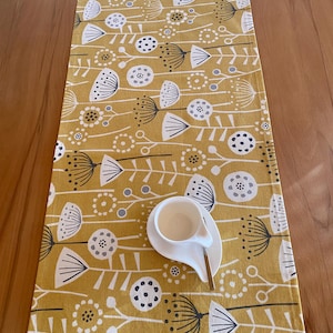 Può includere: Runner da tavolo giallo senape con un motivo floreale bianco, grigio e nero. Una tazza e un piattino bianchi con un cucchiaio poggiano sul runner. Il runner è rettangolare e probabilmente in tessuto.
