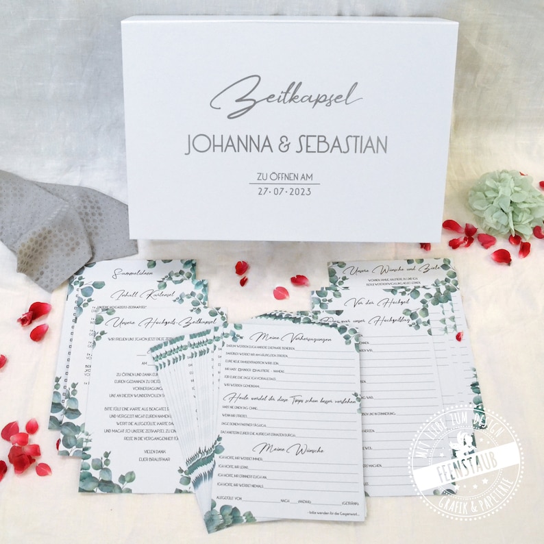 May include: A white box with the text "Zeitkapsel" and "JOHANNA & SEBASTIAN" on the front. The box is surrounded by cards with green eucalyptus leaves and text in German. The text on the box says "ZU OFFNEN AM 27-07-2023".
