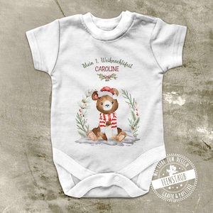 Può includere: Body bianco per neonato con un orso marrone che indossa un cappello da Babbo Natale rosso e una sciarpa. Il body ha la scritta "Mein 1. Weihnachtsfest CAROLINE" stampata.