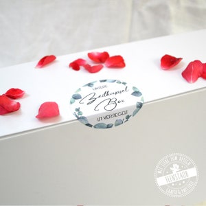May include: White box with a round sticker that says "Unsere Zeitkapsel Box ist versiegelt" in a green leafy design. The box is surrounded by pink rose petals.