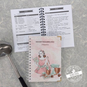 Op de afbeelding: Een spiraalgebonden receptenboek met de titel REZEPTESAMMLUNG Johanna en een roze kaft met een illustratie van iemand die bakt. De open pagina's tonen een handgeschreven Tiramisu-recept in het Duits, inclusief ingrediënten en instructies. Een metalen soeplepel is zichtbaar.