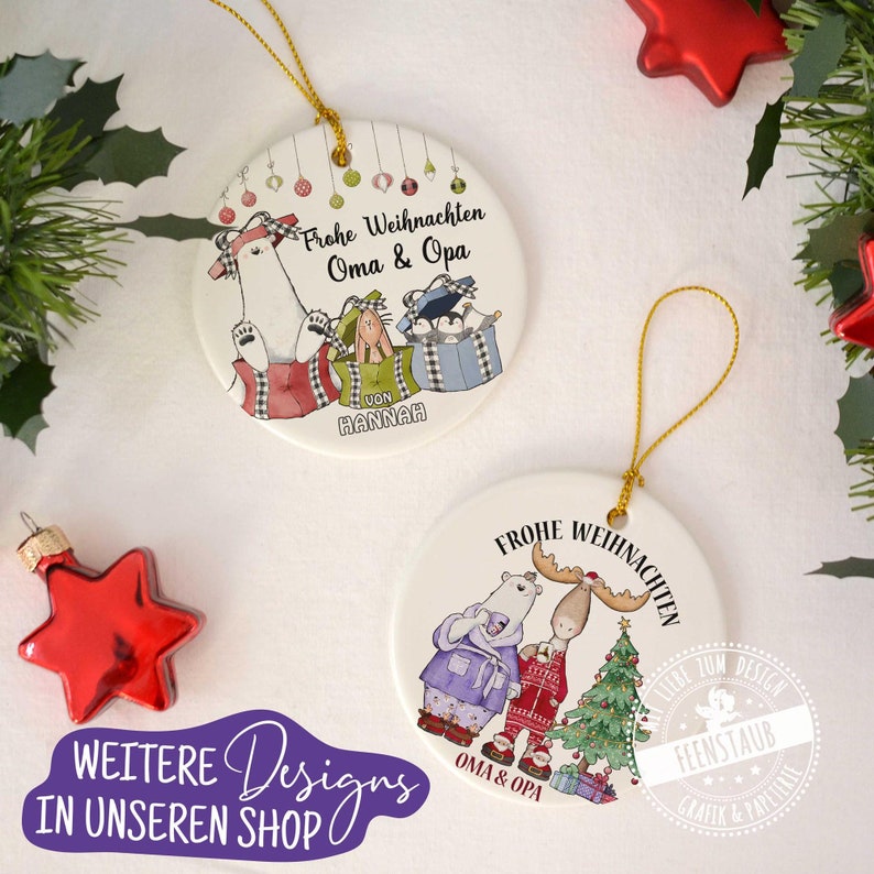 May include: Two white ceramic Christmas ornaments with gold string hangers. The first ornament features a polar bear, rabbit, and penguin holding gifts with the text "Frohe Weihnachten Oma & Opa" and "von Hannah". The second ornament features a polar bear and moose standing by a Christmas tree with the text "Frohe Weihnachten Oma & Opa" and "Liebe zum Design Feenstaub Grafik & Papeterie".