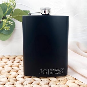 Könnte beinhalten: Eine schwarze Flasche mit silbernem Verschluss und Scharnier. Der Text auf der Flasche lautet "JG TRAUZEUGE 25.10.2025". Die Flasche ist rechteckig. Der Hintergrund ist neutral mit etwas Grün.